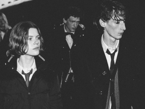 Models wearing tailored dark outerwear with white collared shirts and ties in a black and white shot for Thom Browne.