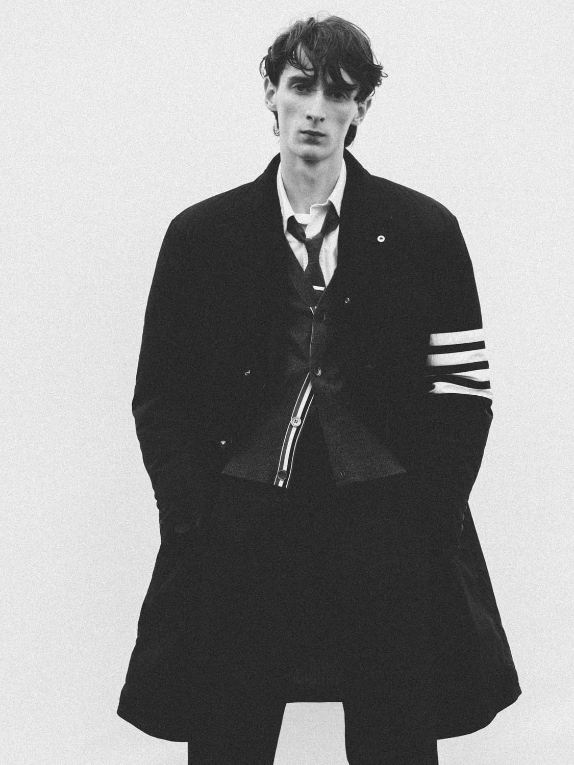 Arthur del Beato wearing a black Thom Browne four-bar overcoat, collared shirt, and tie against a white studio backdrop.
