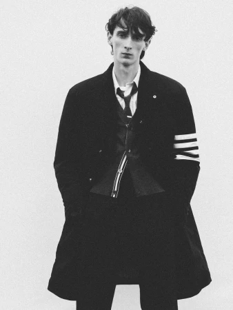 Arthur del Beato wearing a black Thom Browne four-bar overcoat, collared shirt, and tie against a white studio backdrop.