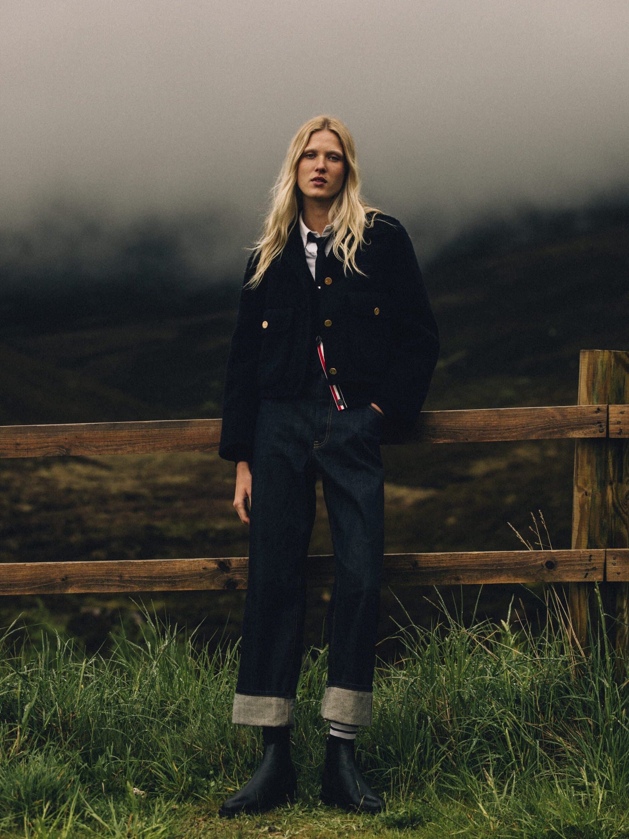 Model standing by a wooden fence in a misty landscape wearing a black jacket and cuffed dark denim trousers.