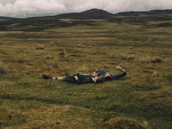 Two men in dark tailored suits and shorts lying together on a wide, grassy moorland under an overcast sky.