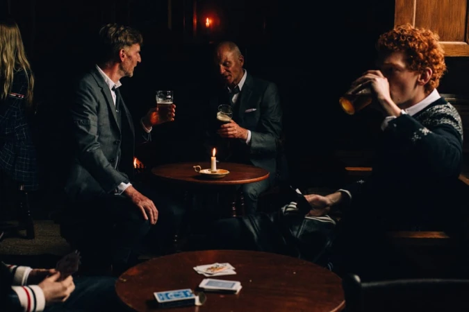 Men wearing tailored suits and knitwear drinking beer around candlelit wooden tables in a dark pub setting.