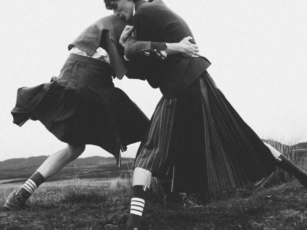 Two male models grappling outdoors on a grassy hillside wearing tailored pleated kilts, blazers, and striped crew socks.