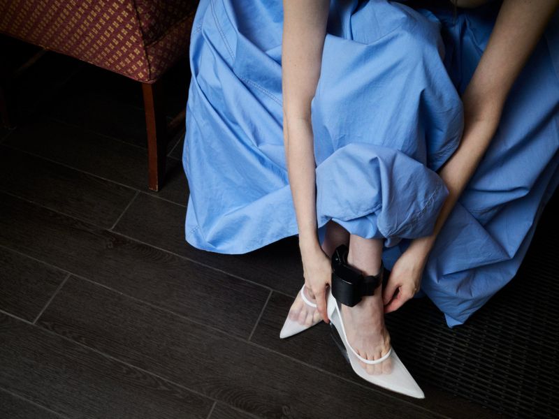 A woman in a blue dress sits adjusting the strap of her white pointed-toe high-heeled shoe, with a black ankle monitor visible.