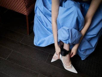 Woman in bright blue dress seated on a dark wooden floor wearing white heels and an ankle monitor
