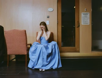 Woman in a long blue dress sitting in a wooden interior with hands covering her face, photographed for Sleek Magazine.
