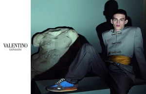 Model in a grey embellished jacket, gold sash, and sneakers seated next to a classical marble torso for Valentino.