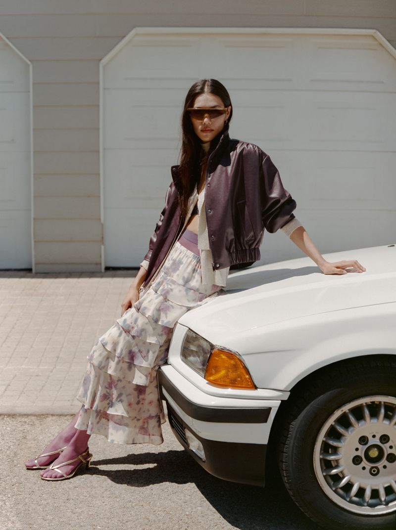 Model Jade Nguyen leaning against the hood of a white car wearing a dark leather jacket, sunglasses, and a ruffled floral skirt.