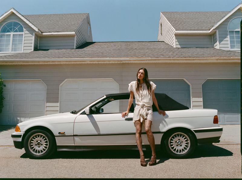Model Jade Nguyen leaning against a white convertible car parked in front of suburban houses under direct daylight.