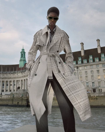 Model in sunglasses wearing a belted plaid trench coat and dark trousers standing outdoors by a river in front of a historic building.