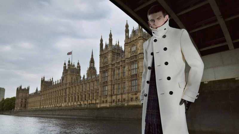 A man in an off-white double-breasted overcoat stands by the River Thames with the Palace of Westminster in the background.
