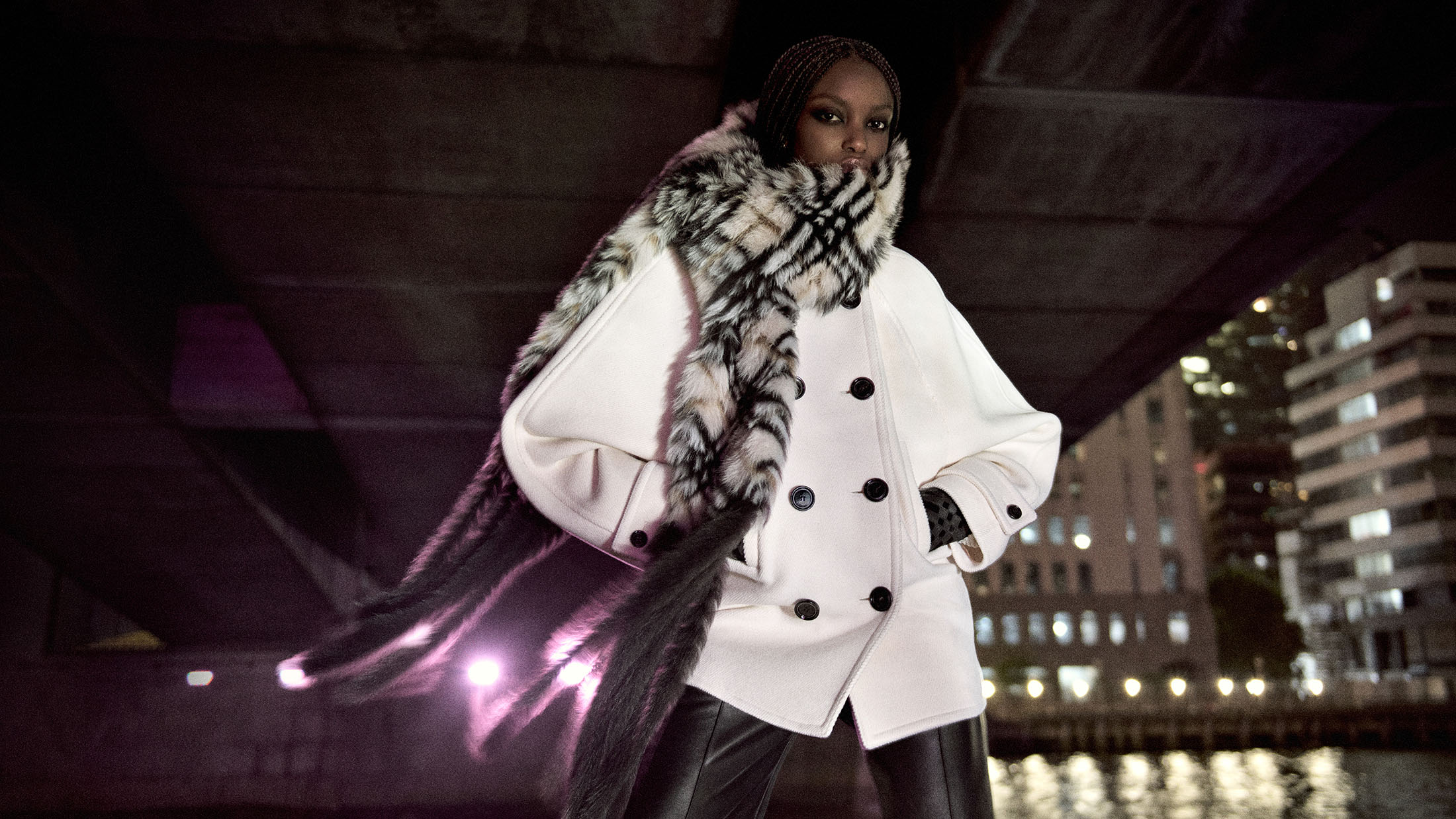 Model posing in an oversized white double-breasted coat with a patterned long fur scarf under a bridge at night.