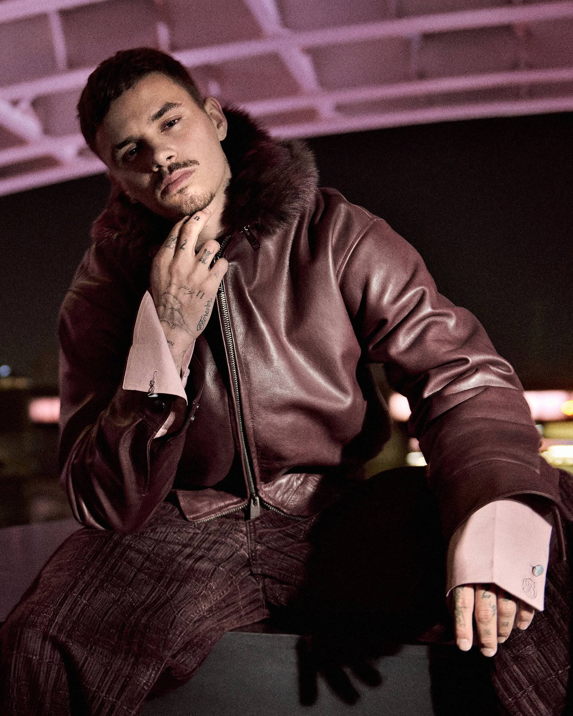 Man in a burgundy leather jacket with a fur collar and pink cuffs sitting outdoors at night.