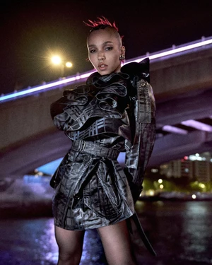 FKA twigs wearing a structured ruffled black mini trench coat outdoors under an illuminated bridge at night.