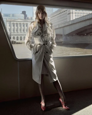 Model posing in an off-white belted ruffled trench coat and red heels before a boat window overlooking a city bridge and river.
