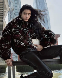Model perched on a metal beam wearing a dark plaid faux-fur jacket and black leather trousers against an urban river skyline.
