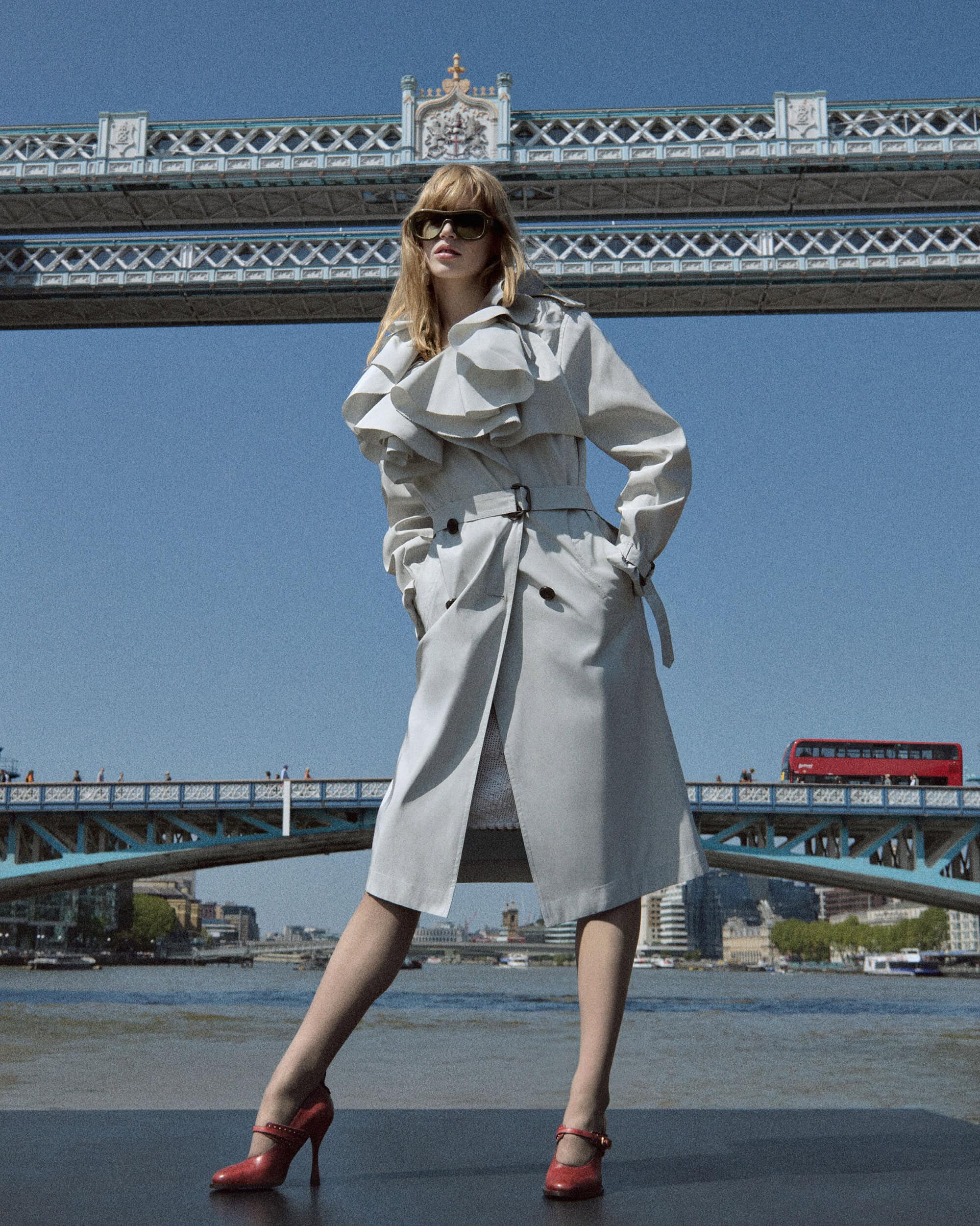 Model posing in an oversized ruffled trench coat, dark sunglasses, and red heels with London's Tower Bridge in the background.