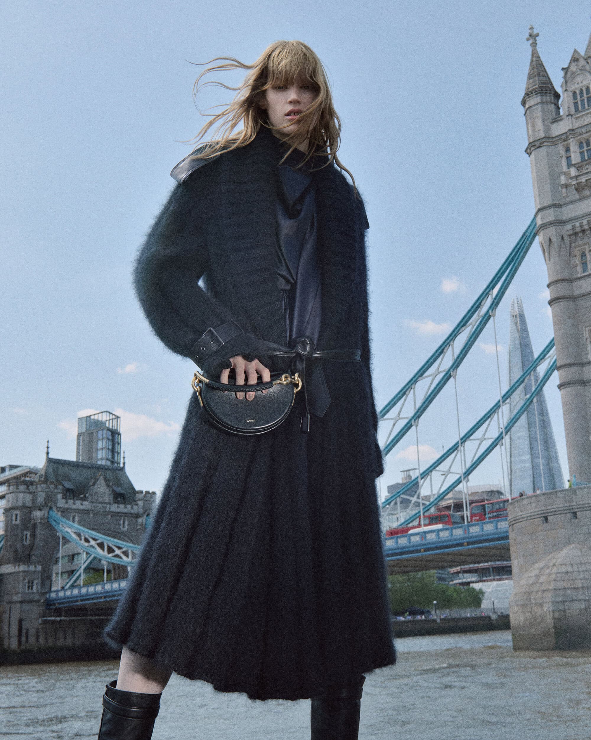 Model with blonde hair wearing a black textured knit coat and boots holding a black crescent handbag against London's Tower Bridge.