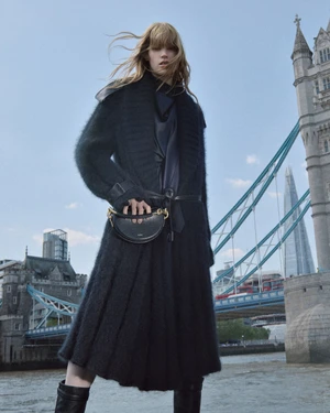 Model with blonde hair wearing a black textured knit coat and boots holding a black crescent handbag against London's Tower Bridge.
