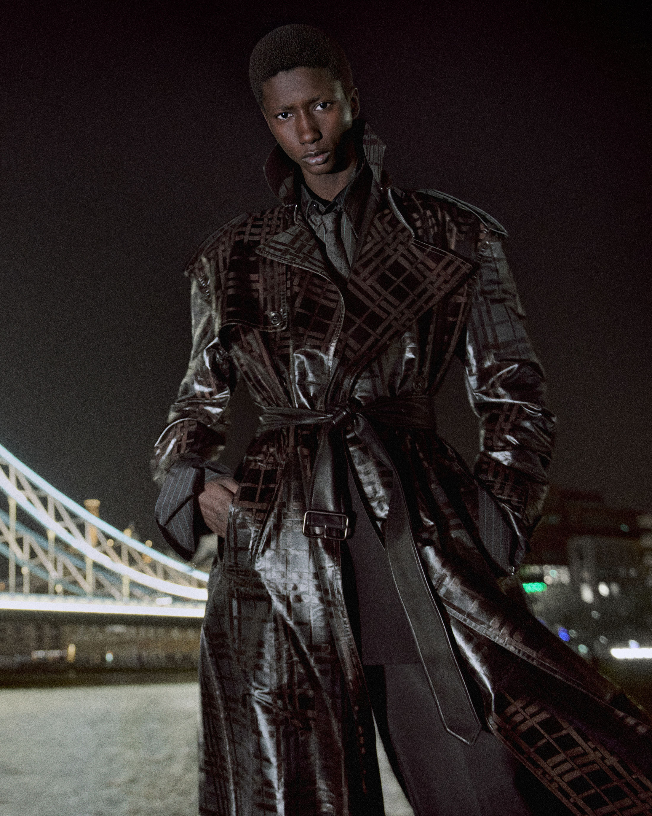 Model wearing a glossy dark check trench coat standing at night in front of an illuminated bridge.