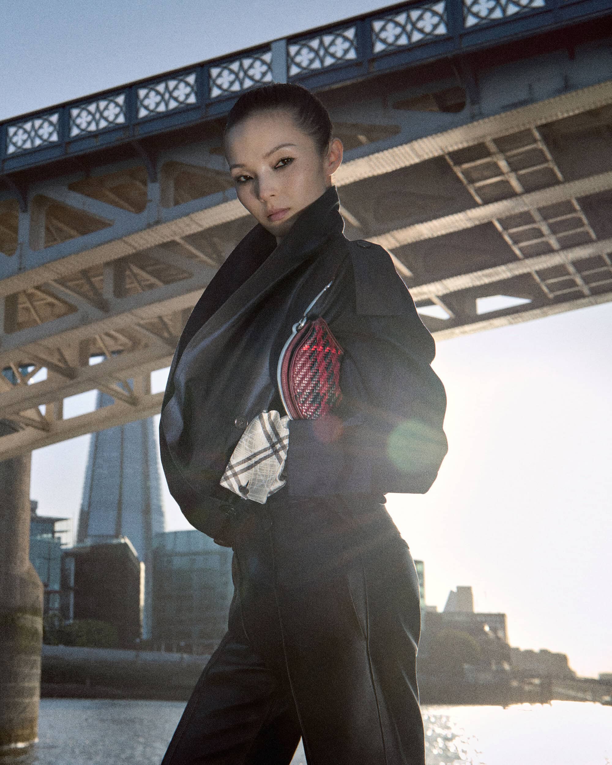 Model Xiao Wen Ju posing in a structured black jacket and trousers beneath an urban bridge overlooking a river.