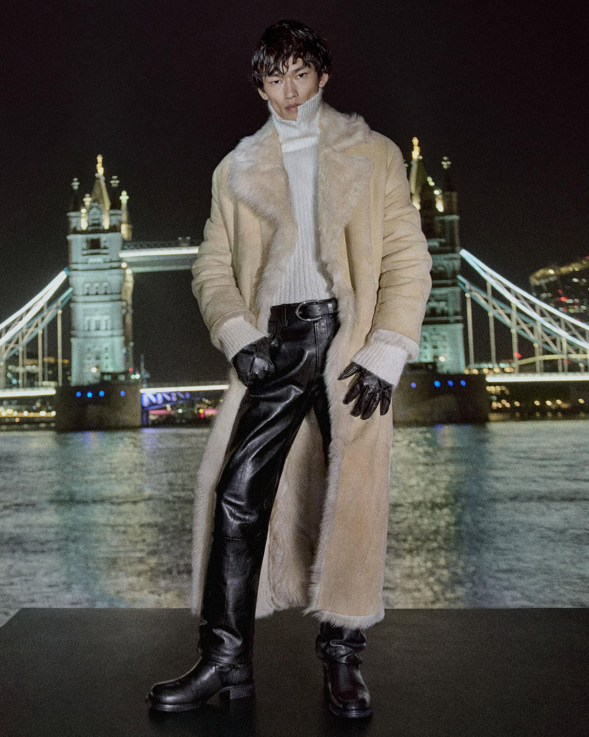 Model in a long beige shearling coat and black leather pants posing at night with London's illuminated Tower Bridge in the background.