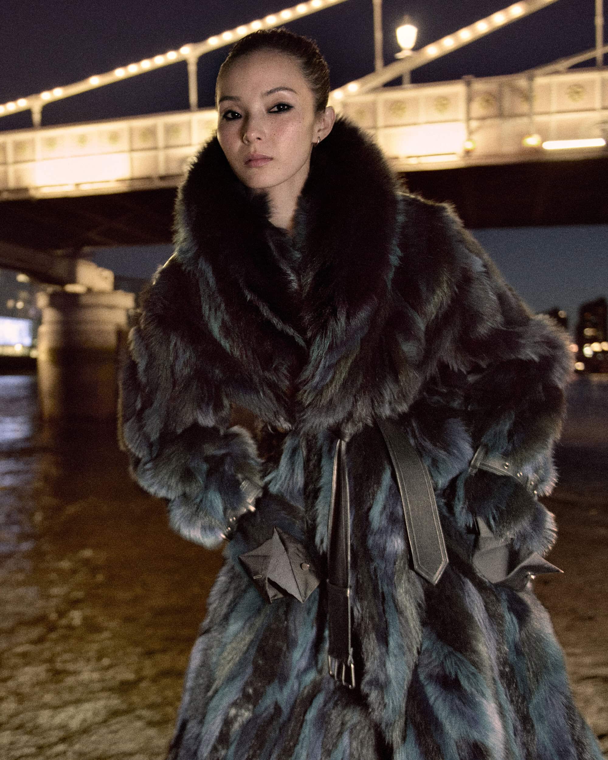 Model Xiao Wen Ju wearing a voluminous dark fur coat in front of an illuminated bridge at night.