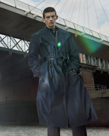 A man wearing a belted navy trench coat stands outdoors beneath the structure of a large suspension bridge.