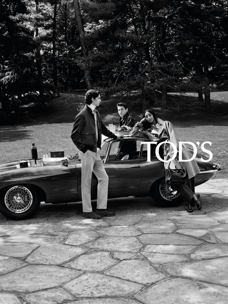 Black and white fashion campaign photograph with three models posing around a vintage sports car on a stone patio surrounded by trees.