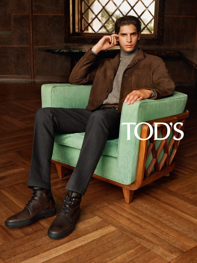 Model wearing a brown suede jacket, grey turtleneck, and trousers seated in a light green armchair on parquet wood flooring.