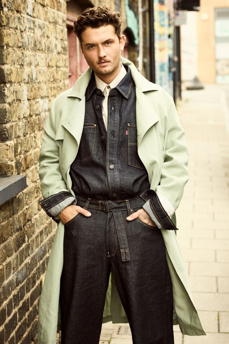 Handsome man in a denim jumpsuit with a trench coat, standing in a brick-lined alley, looking at the camera with a slight smirk.