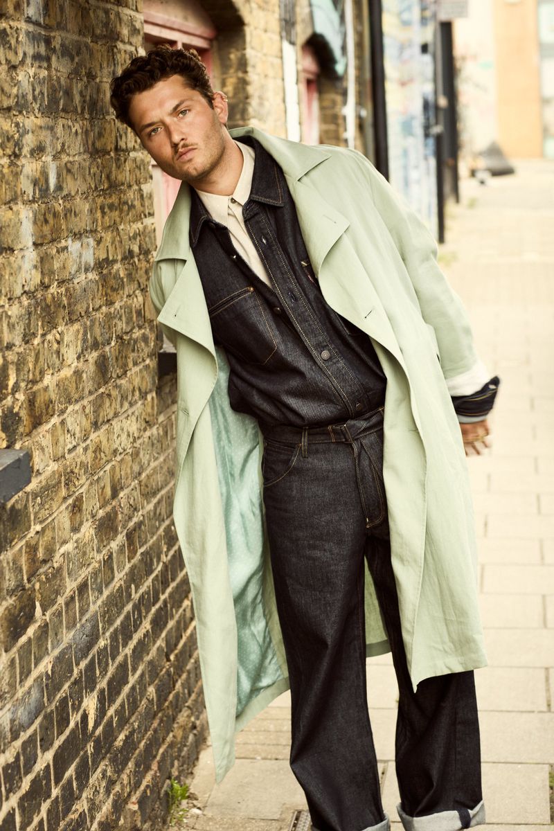 Raff Law leaning against an exterior brick wall wearing a pale green trench coat over a dark denim shirt and matching cuffed jeans.