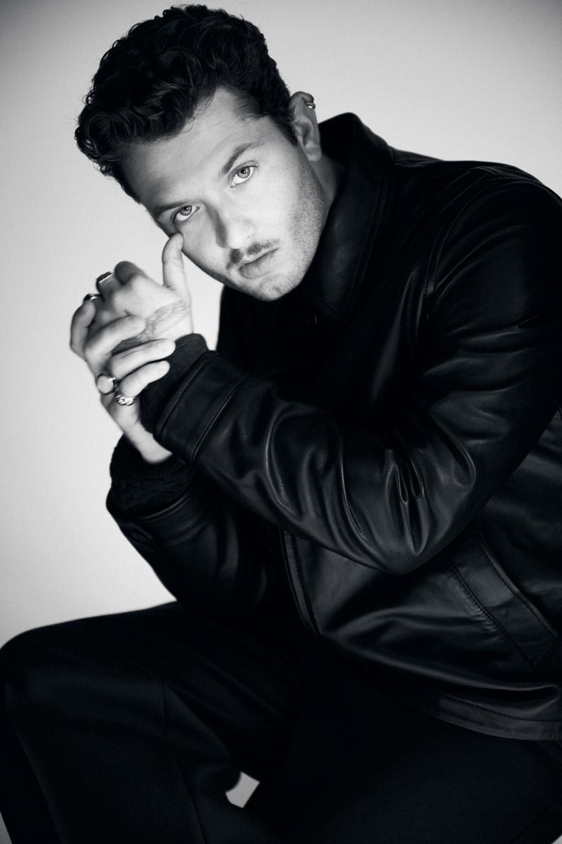 Monochrome portrait of an adult man with dark hair wearing a leather jacket, looking at the camera in a photo studio setting.