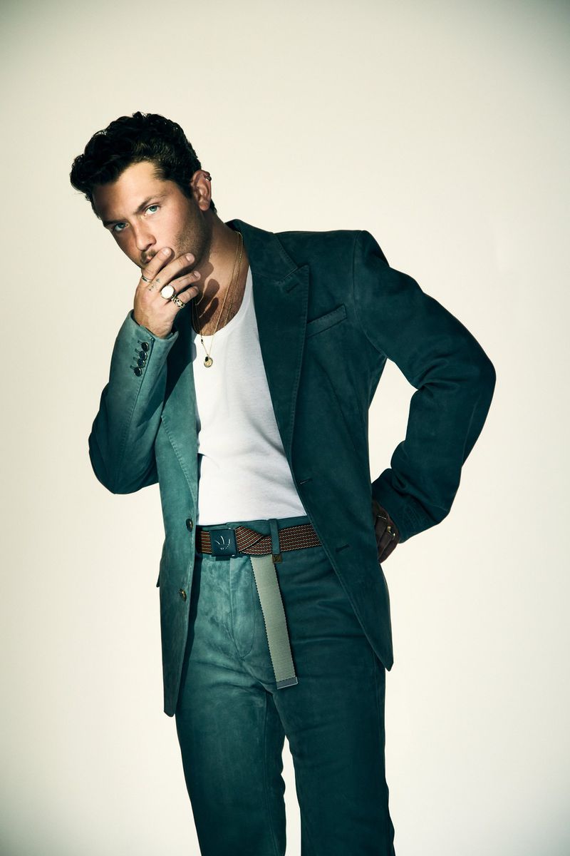 Raff Law posing in a dark green tailored suit jacket, trousers, and a white tank top against a pale studio backdrop.