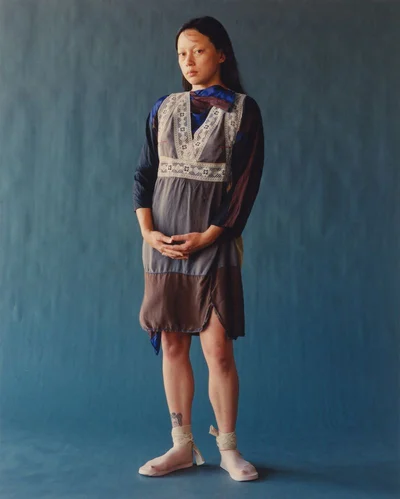 East Asian woman in a layered panel dress and lace ankle wrap shoes posing against a deep blue studio wall.