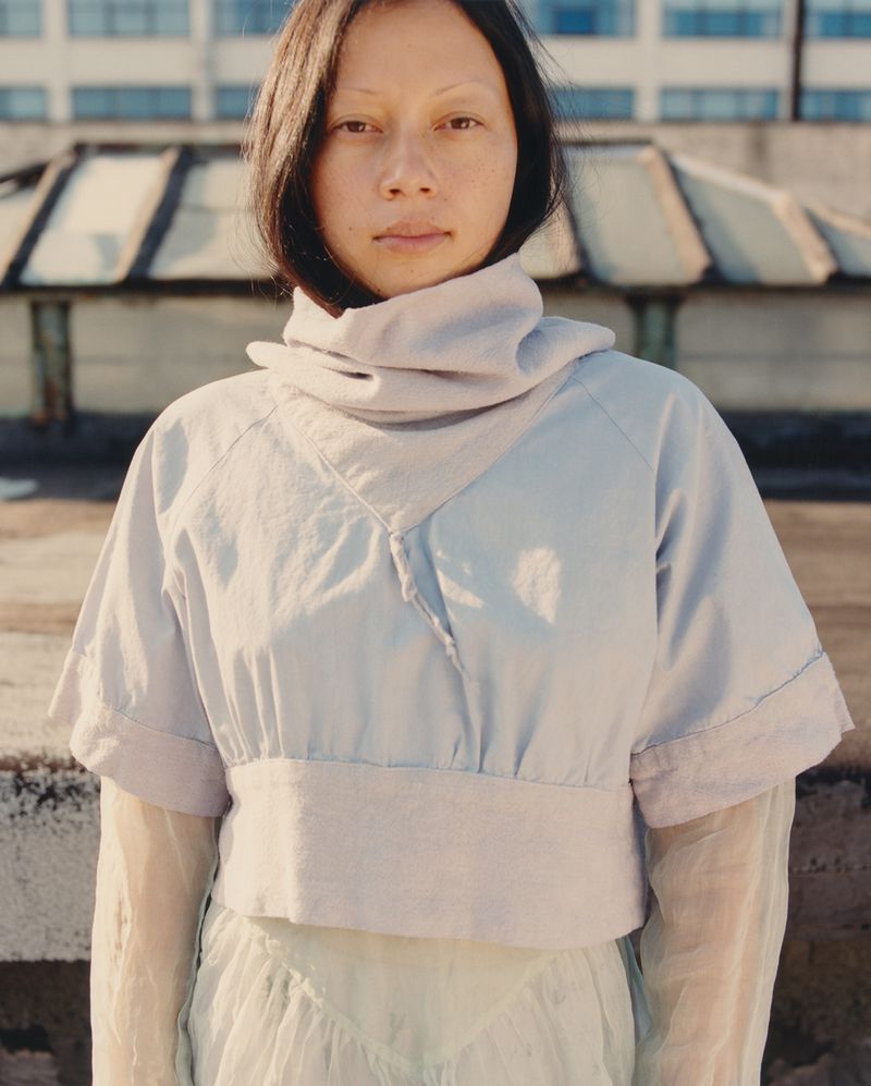 An East Asian woman wearing a high-collared light blue cropped top standing on an urban rooftop with industrial skylights behind her.