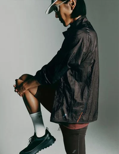 Model Hyuk Kim in a dark performance jacket, cap, and running shoes stretching against a studio background.