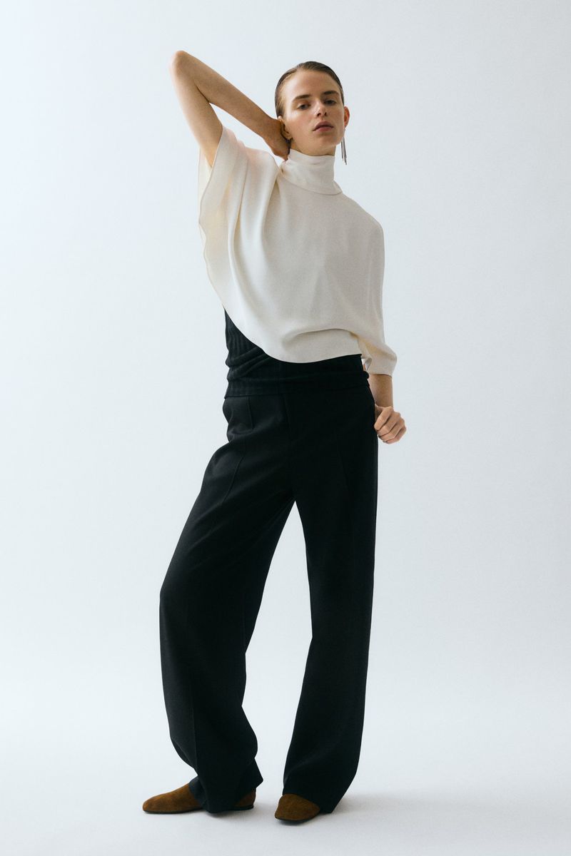 Model posing in an off-white high-neck draped blouse and wide-leg black trousers against a seamless white studio backdrop.
