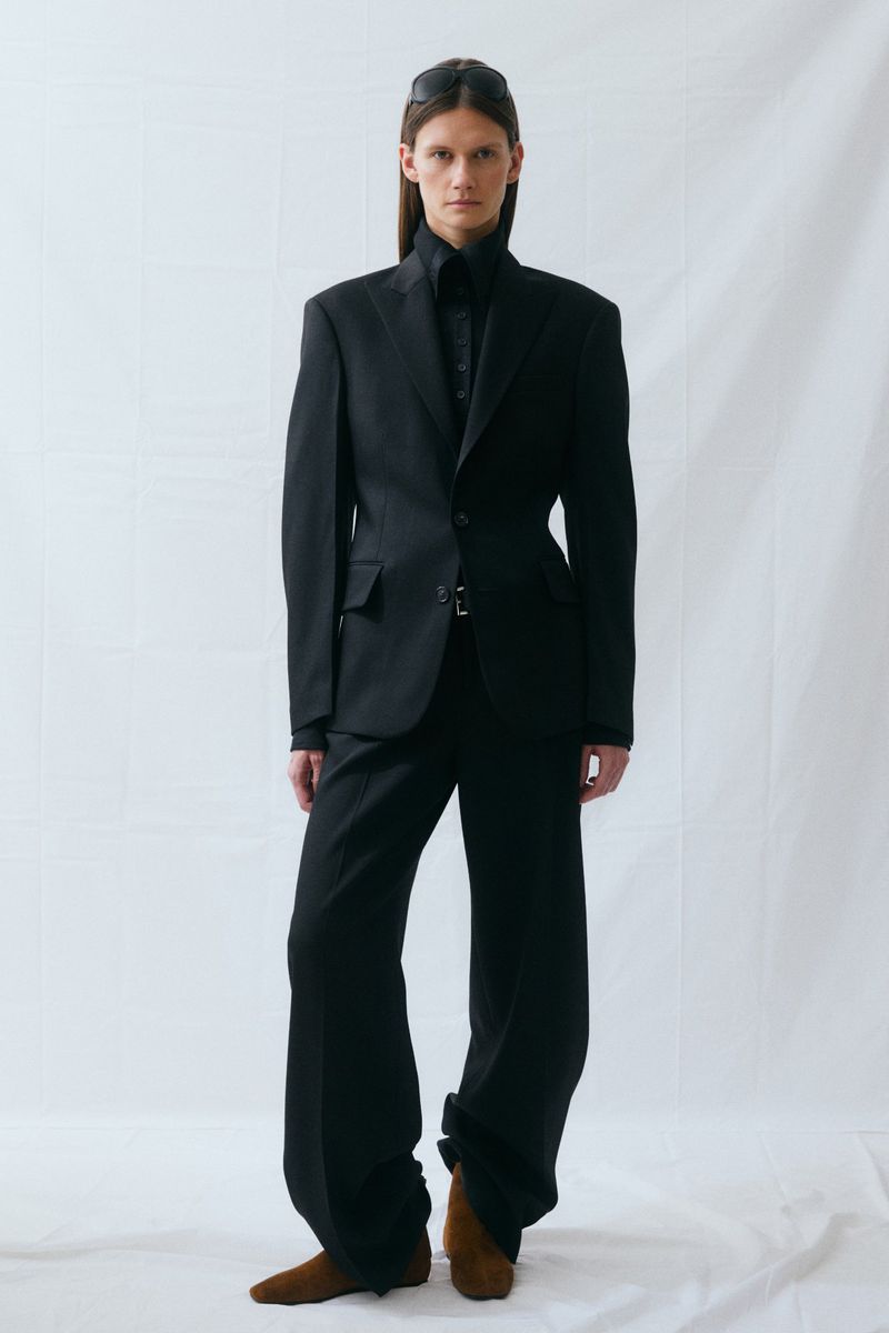 Fashion model wearing a sharp black tailored suit blazer, wide-leg trousers, and brown suede shoes against a white backdrop.
