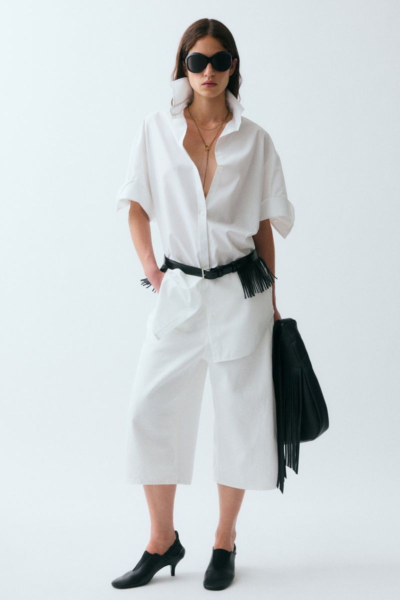 Model wearing an oversized white shirt, white culottes, black sunglasses, and black fringed accessories against a white background.