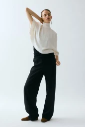 Model posing in a draped cream high-neck top and wide-leg black trousers with brown flats on a white studio background.