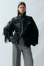 Model wearing a black fringe leather jacket and grey tailored trousers against a white studio backdrop.
