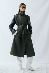 Model wearing dark sunglasses and a charcoal belted trench coat with black sleeves over white pants in front of a white backdrop.