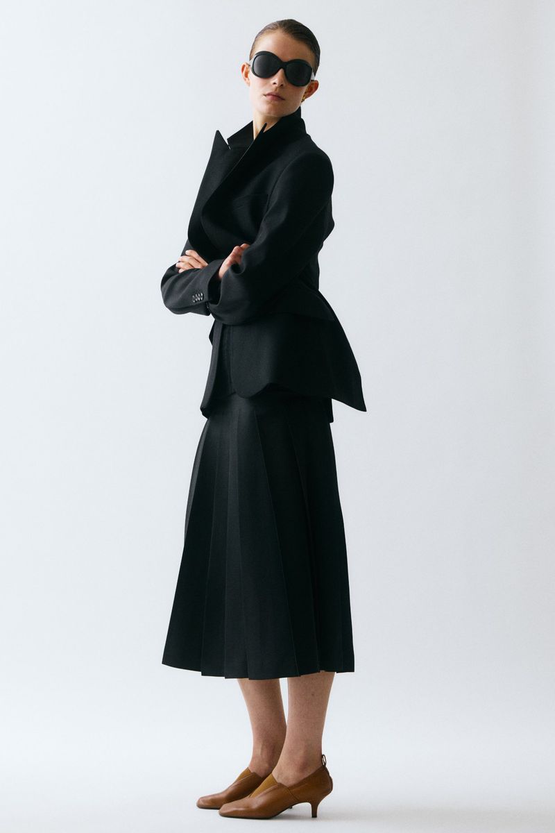 Model wearing a black tailored peplum jacket, pleated midi skirt, dark sunglasses, and tan kitten heels against a white backdrop.