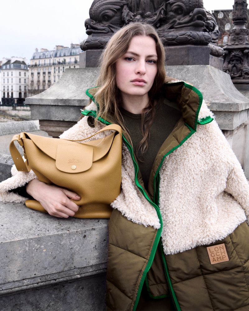 Model Felice Nova Noordhoff holding a mustard leather shoulder bag while wearing a fleece and quilted coat on a stone bridge.