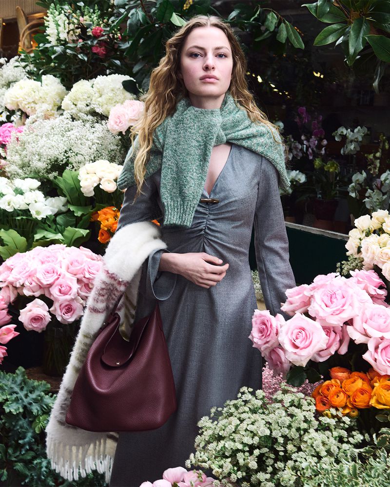 Felice Nova Noordhoff in a grey dress and green knit sweater holding a burgundy leather handbag surrounded by pink roses and flowers.