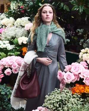 Felice Nova Noordhoff in a grey dress and green knit sweater holding a burgundy leather handbag surrounded by pink roses and flowers.