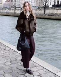 Model Felice Nova Noordhoff in a brown shearling jacket, burgundy trousers, and a black shoulder bag by a riverbank.