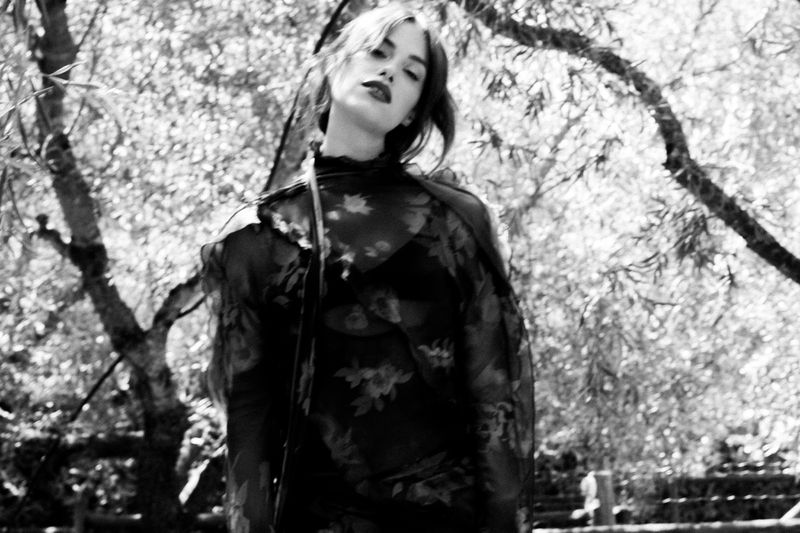 Black and white portrait of a woman in a sheer high-neck floral top outdoors beneath dappled tree branches.