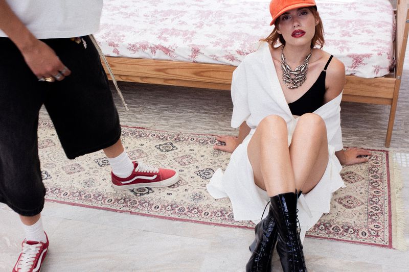 Woman in an orange cap, white wrap, and black lace-up boots sitting on a rug next to a person in black shorts and red sneakers.